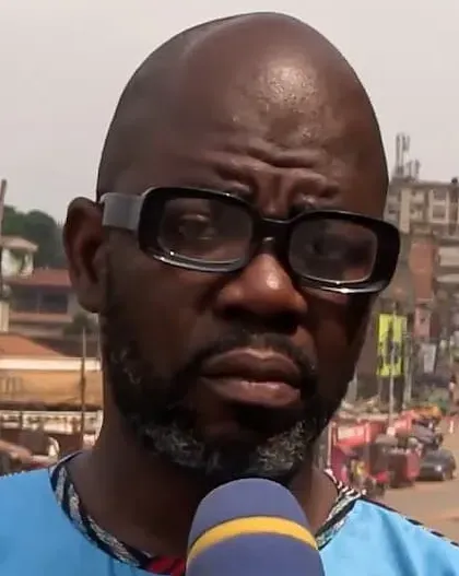 Ernesto Yene, en tunique bleue et lunettes, interviewé face à un micro dans une rue commerçante animée.